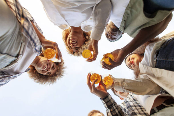 Gruppe von multirassischen jungen Leuten klirrt draußen mit Bierflaschen, interrassische Studenten feiern gemeinsam Toast und entspannen sich, sechs Flaschen Alkohol halten Händchen, Blick von unten - Foto, Bild