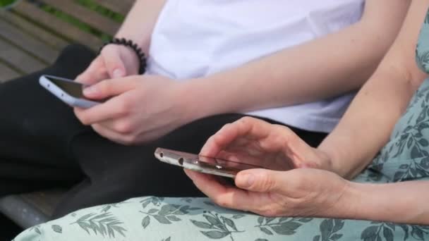 Een volwassen moeder en een tienerzoon zitten op een bankje in een zomerpark en kijken naar een smartphone, drukken op het scherm, houden de telefoon in hun handen. - Video