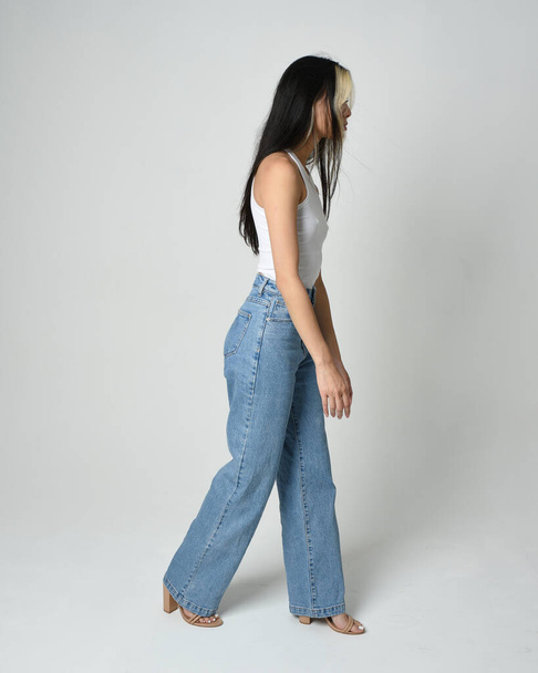 Full length portrait of brunette female model wearing casual clothes, white singlet shirt, denim jean pants. Standing pose walking away from camera. Isolated on white studio background. - Photo, Image