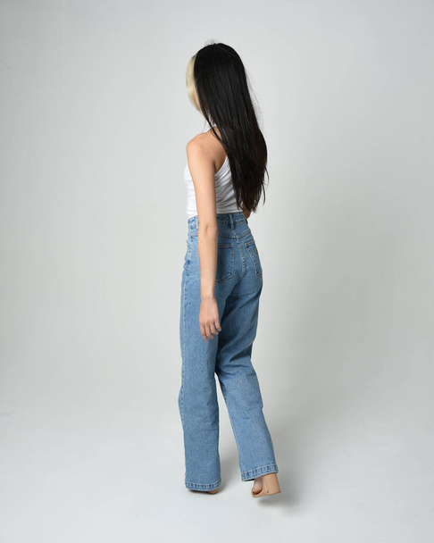 Full length portrait of brunette female model wearing casual clothes, white singlet shirt, denim jean pants. Standing pose walking away from camera. Isolated on white studio background. - Photo, Image