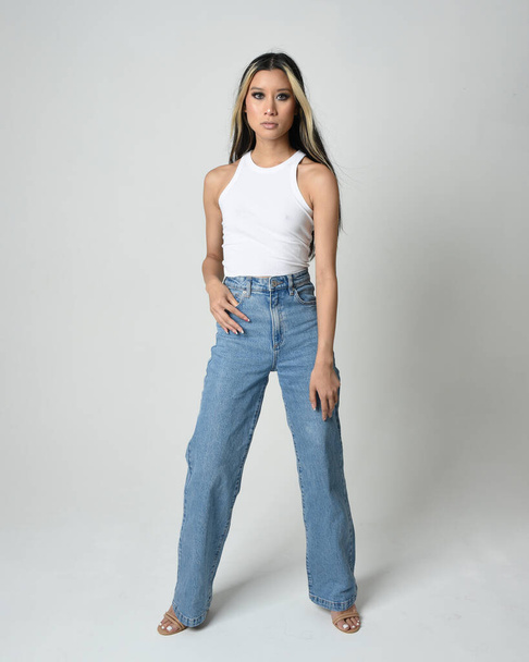Full length portrait of brunette female asian model wearing casual clothes, white singlet shirt, denim jean pants. Standing pose facing towards camera. Isolated on white studio background. - Photo, Image