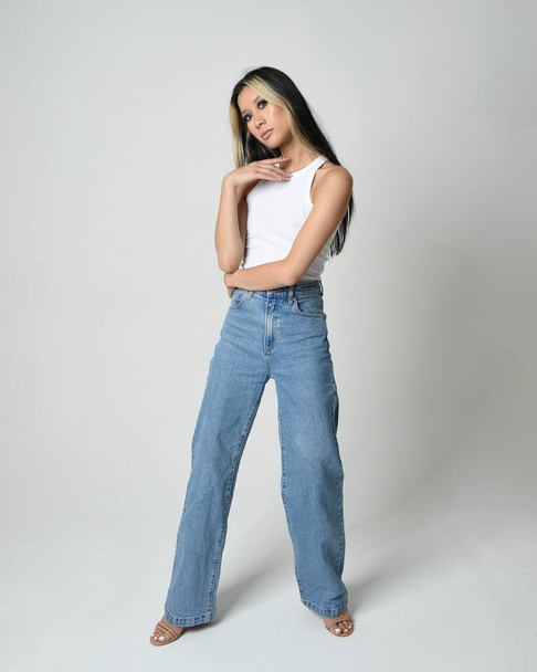 Full length portrait of brunette female asian model wearing casual clothes, white singlet shirt, denim jean pants. Standing pose facing towards camera. Isolated on white studio background. - Photo, Image