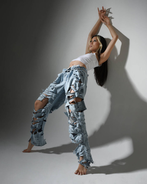 Портрет брюнетки жіночої азіатської моделі в повсякденному одязі, біла сорочка-синглет, джинсові штани. Позиція стоячи, низький кут камери для перспективи. Ізольовані на білому фоні студії. - Фото, зображення