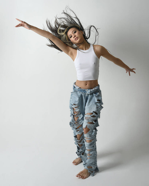 Full length portrait of brunette female asian model wearing casual clothes, white singlet shirt, denim jean pants.  Energetic jumping, dancing pose. Isolated on white studio background. - Photo, Image