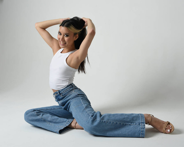 Full length portrait of brunette female asian model wearing casual clothes, white singlet shirt, denim jean pants. Sitting pose, high camera angle for perspective. Isolated on white studio background. - Foto, Imagem