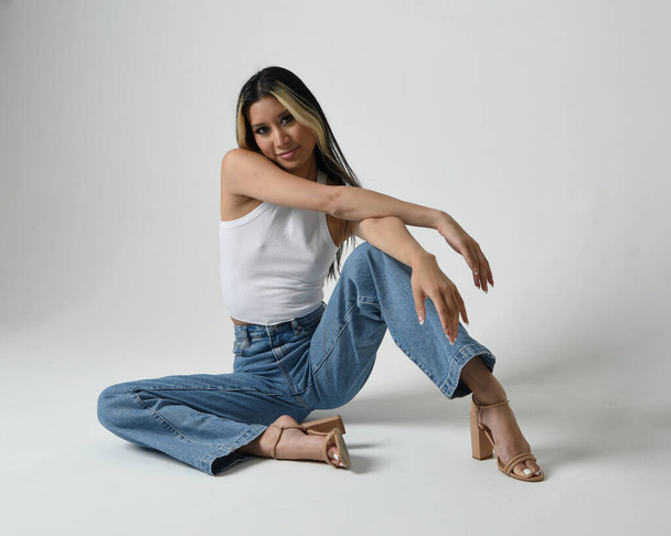 Full length portrait of brunette female asian model wearing casual clothes, white singlet shirt, denim jean pants. Sitting pose, high camera angle for perspective. Isolated on white studio background. - Photo, Image