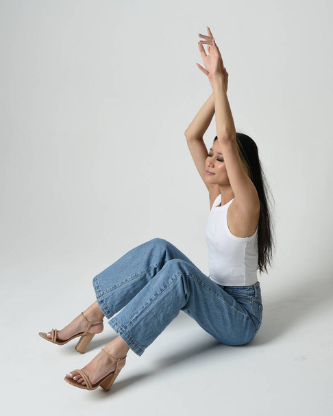 Full length portrait of brunette female asian model wearing casual clothes, white singlet shirt, denim jean pants. Sitting pose, high camera angle for perspective. Isolated on white studio background. - Foto, immagini