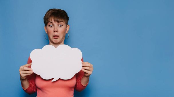 Foto de estudio de una joven mujer con una expresión facial extraña, sosteniendo un estandarte blanco vacío en forma de nube sobre un fondo azul. Copia espacio, espacio para tu anuncio o texto - Foto, imagen
