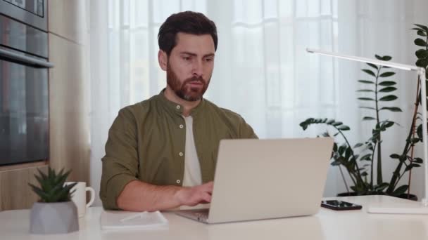 大人の男性はポータブルコンピュータで机に座って,思慮深く遠ざかっている間に電子メールを書いています. 生産的な作業プロセスのための軽いアパートで居心地の良い国内オフィスを選択するコーカサスフリーランサー. - 映像、動画