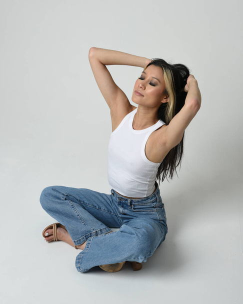 Pełna długość portret brunetka kobieta azjatycki model noszenie casual ubrania, biały koszulka singlet, jeansowe spodnie jeansowe. Siedząca pozycja, duży kąt widzenia kamery. Izolacja na białym tle studia. - Zdjęcie, obraz