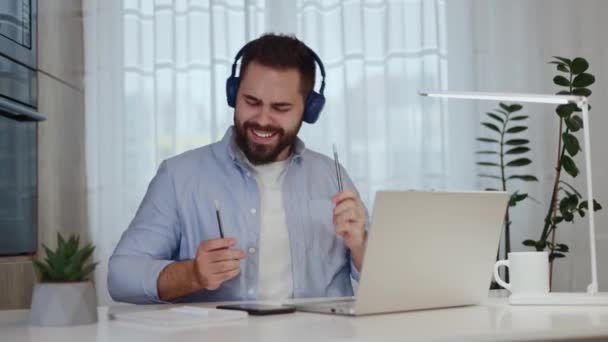 Vidám távoli dolgozó kék fülhallgatóval okostelefont használ a zeneváltáshoz és a ritmusmozdulatokkal való zenéléshez. Gadget felhasználó tesztelés új mobil alkalmazás, miközben ül a hazai munkaterület. - Felvétel, videó