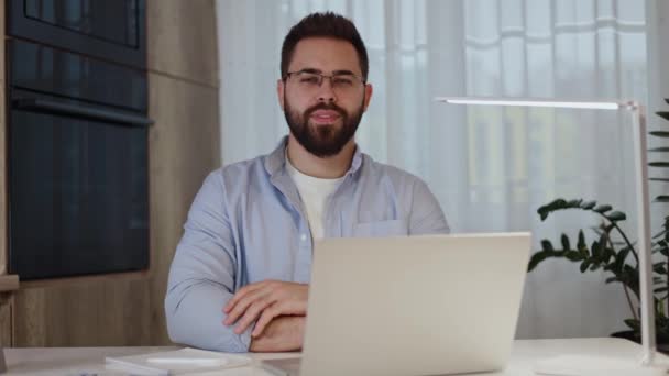 Satisfied attractive man looking at camera with smile and crossed arms while sitting by laptop at home. Successful freelance designer having break during working day and enjoying flexible schedule. - Footage, Video