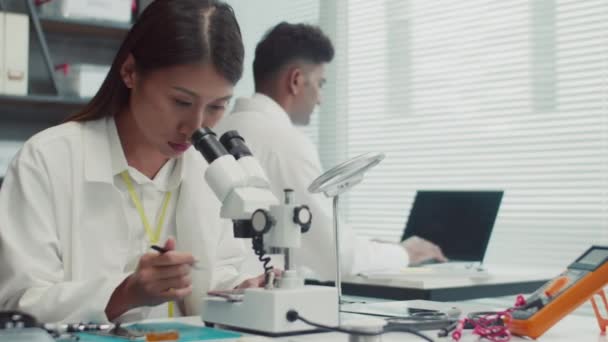 Colpo medio di tecnico donna in labcoat bianco guardando al microscopio quando si fissa componente smartphone rotto al centro di riparazione - Filmati, video