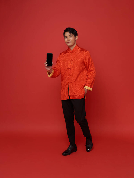 Onnellinen aasialainen mies yllään perinteinen mekko osoittaa matkapuhelimen tyhjä näyttö eristetty punaisella taustalla. - Valokuva, kuva