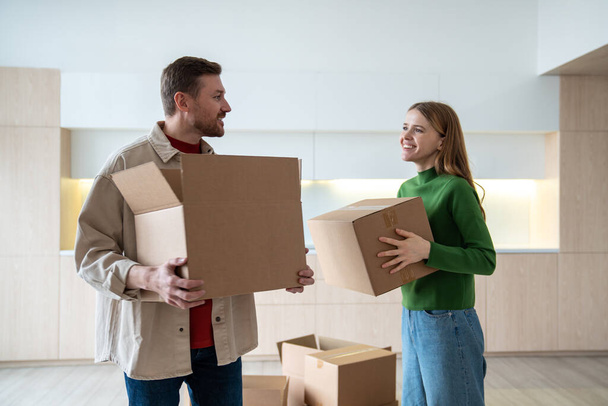 Happy new homeowners tenants renters pleased man and woman carrying boxes to room. Family couple looking eye to eye standing on kitchen while moving to apartment. Relocation, family mortgage concept. - Photo, Image