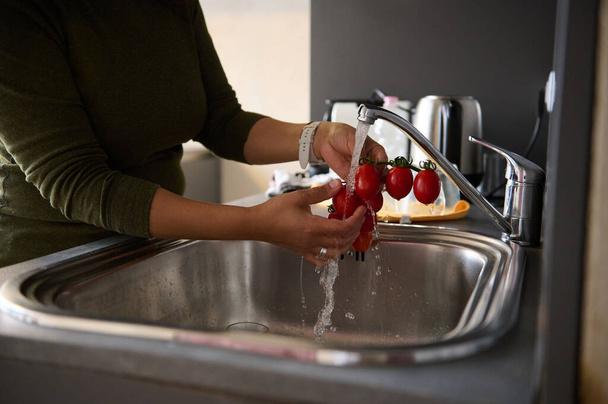 Zbliżenie gospodyni domowej myjącej dojrzałe organiczne wiśnie pomidorowe pod kranem bieżącą wodą, w domowej kuchni - Zdjęcie, obraz