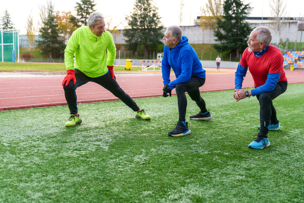 Un grupo de atletas de edad avanzada en ropa deportiva participan en estiramientos laterales en una pista de atletismo, mostrando el envejecimiento activo - Foto, imagen