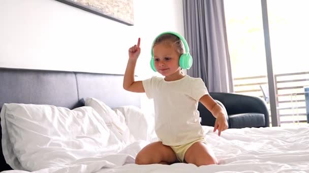 Kislány élvezi a zenét a gyerekek fejhallgató és tánc az ágyban otthon. Kiváló minőségű 4k felvételek - Felvétel, videó