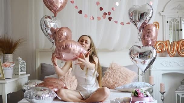 Beautiful young girl at home on the bed in the morning enjoy valentines day celebration with heart shaped balloons. High quality 4k footage - Metraje, vídeo