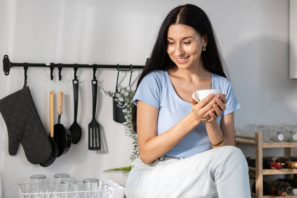 Hermosa joven mujer bebiendo mañana sosteniendo taza de café sentado en el mostrador de cocina moderna disfrutando de relajación.  - Foto, imagen