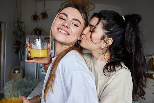 Uczuciowa para lesbijek po dwudziestce dzieląca się pocałunkiem, kobieta trzymająca świeży sok w przytulnej kuchni - Zdjęcie, obraz