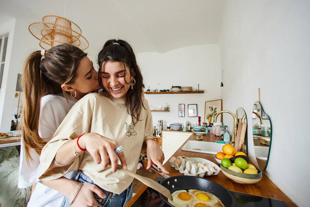 happy and young lesbian couple in 20s cooking breakfast and kissing in kitchen, intimate moment - Photo, Image
