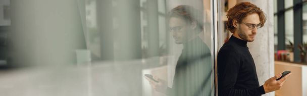 Красивий чоловічий фрілансер тримає телефон, стоячи в офісі під час перерви - Фото, зображення