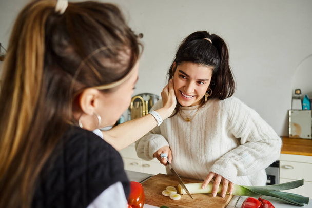 lgbt pärchen, junge lesbische frau beim haaranpassen ihrer glücklichen freundin beim schnitzen von porree in der küche - Foto, Bild