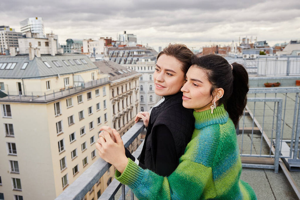 Neşeli lezbiyen çift, gündelik giysiler içinde çatıdaki şehre bakıyor, bir anlık aşk ve bağ. - Fotoğraf, Görsel