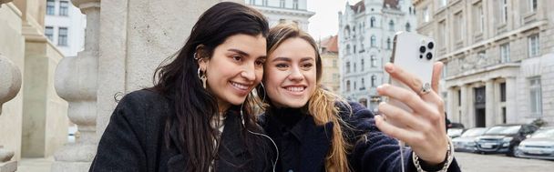 ウィーンの屋外で一緒に座っている間,スマートフォンでセルフィーを取る幸せなレズビアンカップルのバナー - 写真・画像