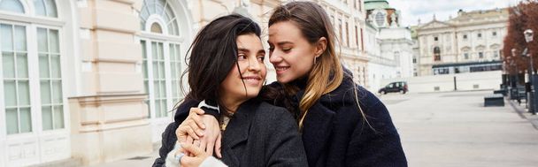 glückliches lesbisches Paar in Oberbekleidung lächelnd, während es sich in der Nähe des Gebäudes in Wien umarmt, Banner - Foto, Bild