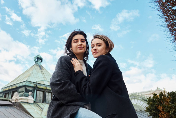 wesoła para lesbijek w odzieży wierzchniej uśmiechająca się podczas przytulania w Wiedniu, patrząc w kamerę - Zdjęcie, obraz