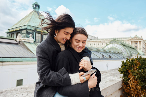 happy lesbian woman holding smartphone and hugging her girlfriend during trip to Vienna - Photo, Image