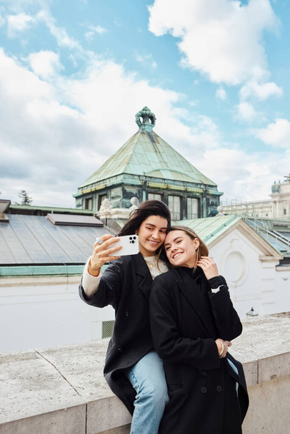 szczęśliwy lgbt para biorąc selfie na smartfonie stojąc razem na ulicy w Wiedniu, lesbijki - Zdjęcie, obraz