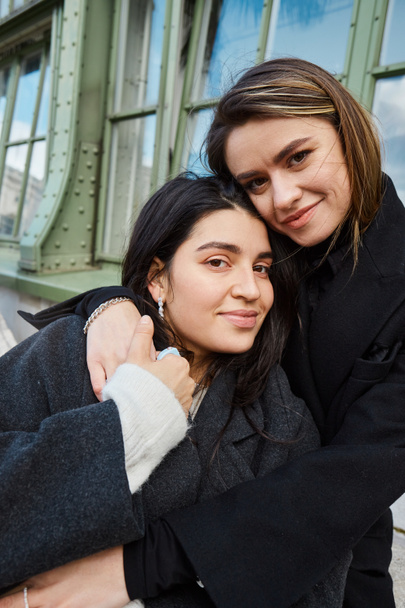 portrait of cheerful lesbian couple in coats embracing each other near Palmenhaus in Vienna - Photo, Image