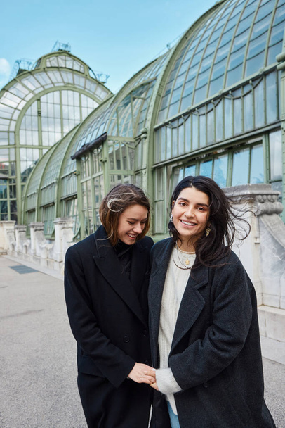lgbt casal em casacos da moda preto de mãos dadas enquanto ri perto de Palmenhaus em Viena - Foto, Imagem