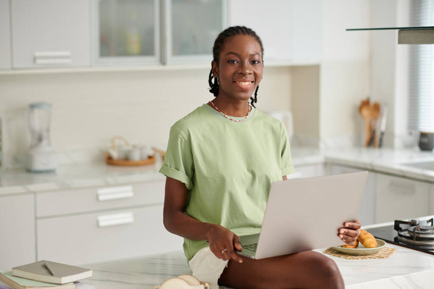 Portrét veselé černošky dívka s notebookem sedí na ostrově kuchyně - Fotografie, Obrázek