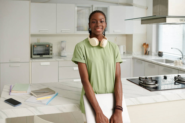 Весела чорна дівчина з навушниками та ноутбуком стоїть на кухонній стійці - Фото, зображення