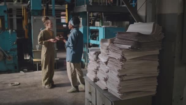 Colpo completo di due impiegate multirazziali in abbigliamento da lavoro che conversano durante il turno di lavoro presso la stamperia con pile di carta sul vecchio cassetto in primo piano focalizzato - Filmati, video