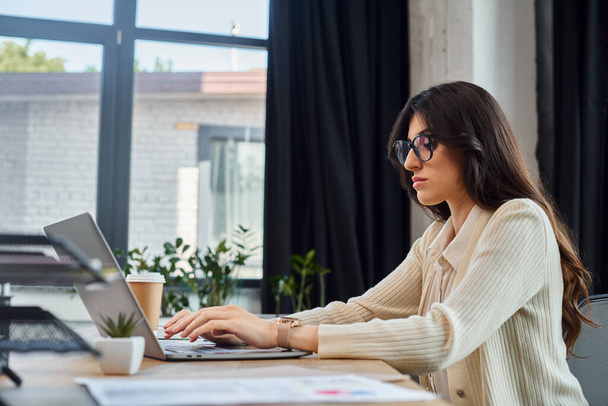 Una mujer de negocios se sienta en un escritorio, centrado en la pantalla de su computadora portátil, en un entorno de oficina moderno con artículos relacionados con la franquicia. - Foto, Imagen