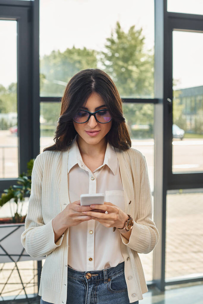 Une femme d'affaires concentrée se tient près d'une fenêtre, vérifiant son téléphone portable dans un cadre de bureau moderne. - Photo, image