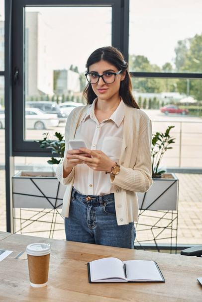 Μια εστιασμένη επιχειρηματίας στέκεται σε ένα τραπέζι σε ένα σύγχρονο γραφείο, βαθιά απορροφημένη στο τηλέφωνό της, σκεπτόμενη ιδέες Franchise. - Φωτογραφία, εικόνα