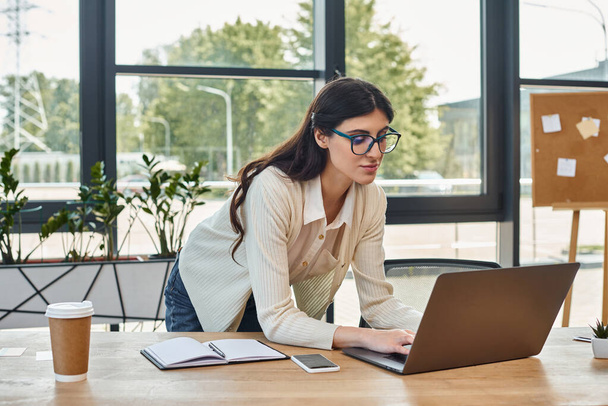Una mujer de negocios está sentada en una mesa, enfocada en su computadora portátil en un entorno de oficina moderno, encarnando la esencia del trabajo de franquicia.. - Foto, Imagen