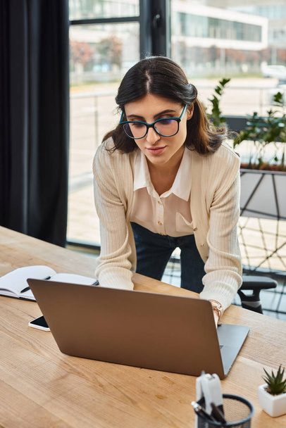 Бізнес-леді в окулярах зосереджена на своєму ноутбуці, керуючи франшизою в сучасному офісному робочому просторі. - Фото, зображення