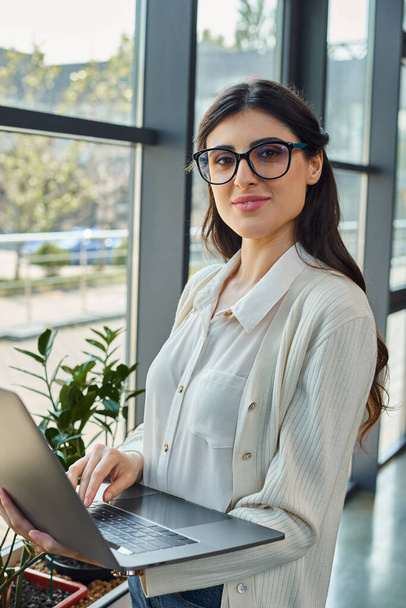 Een gesofisticeerde vrouw met een bril heeft een laptop in een modern kantoor en belichaamt innovatie in de zakenwereld.. - Foto, afbeelding