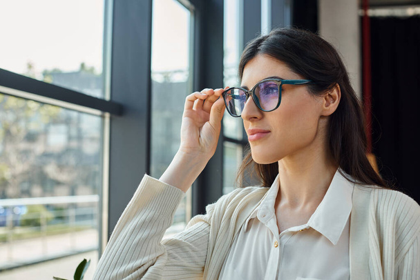 Bizneswoman, w okularach, spogląda przez okno w nowoczesnym biurze, kontemplując miejski krajobraz. - Zdjęcie, obraz