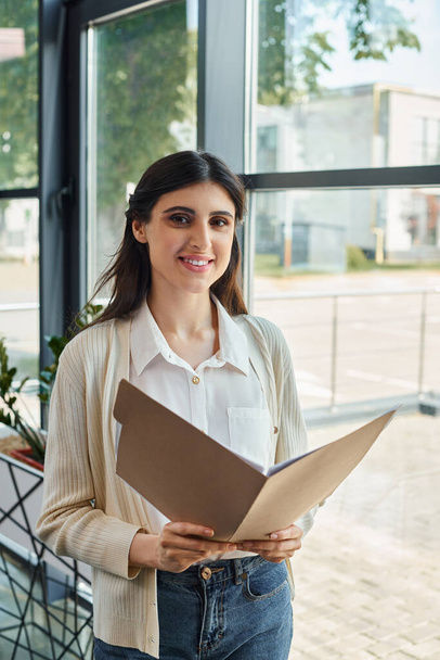 Μια επιχειρηματίας στέκεται με αυτοπεποίθηση μπροστά από ένα παράθυρο, κρατώντας ένα φάκελο, ενσωματώνοντας την ουσία ενός σύγχρονου CEO σε ένα γραφείο Franchise. - Φωτογραφία, εικόνα