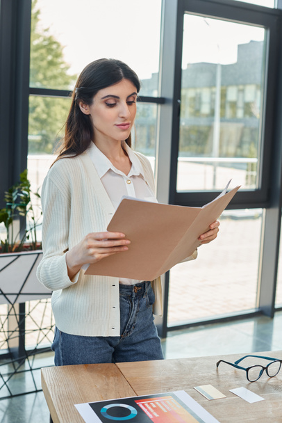 A businesswoman stands in a modern office, holding a piece of paper, with a table in the background hinting at a franchise concept. - Photo, Image