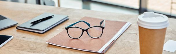 Een bril rust op een levendig notitieboekje naast een stomend kopje koffie, in een moderne kantoorruimte. - Foto, afbeelding