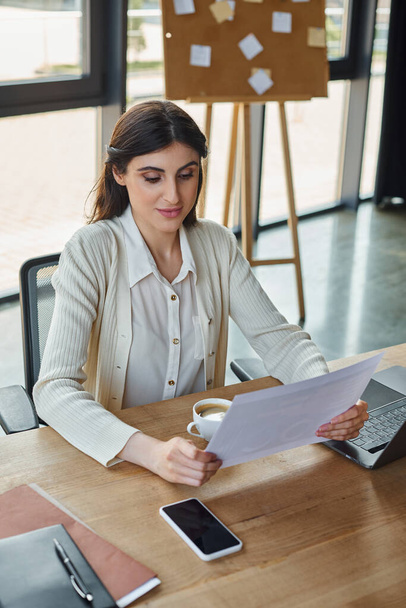 Een moderne zakenvrouw zit aan een tafel en geniet van een moment van ontspanning met een kopje koffie in een chique kantooromgeving. - Foto, afbeelding